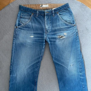 Blå slitna jeansbyxor - Säljer ett par klassiska blå jeansbyxor med slitningar och lätt tvättad look. Byxorna har fem fickor, normal passform och raka ben. Perfekta för en avslappnad stil. Jeansen har en liten reva på bakre fickan, därav är priset väldigt lågt. Det syns inte så mycket men går även lätt att laga!