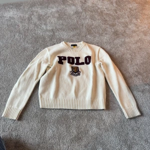 Vit stickad tröja från Polo Ralph Lauren - Säljer en vit stickad tröja från Polo Ralph Lauren med broderad text 'POLO' och ett emblem på bröstet. Tröjan har rund halsringning och långa ärmar. Perfekt för en klassisk och stilren look.