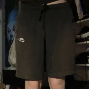 Svarta Nike shorts med snörning - Svarta shorts från Nike med vit logga på vänster ben och snörning i midjan. Modellen är knälång och har en avslappnad passform, perfekt för träning eller chill. Materialet känns mjukt och lätt.