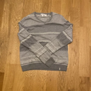 Grå randig tröja från Calvin Klein - Säljer en stilren grå tröja från Calvin Klein i storlek M. Tröjan har ett diskret randigt mönster i olika grå nyanser och v-ringad hals. Perfekt för en avslappnad men snygg look.