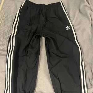 Svarta Adidas byxor med vita ränder - Säljer ett par klassiska svarta Adidas byxor med vita ränder längs sidorna och Adidas-logga på låret. Byxorna har resår i midjan och är perfekta för en sportig stil.