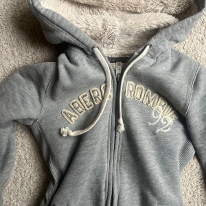 Grå hoodiejacka med teddyfoder från Abercrombie & Fitch - Mysig grå hoodiejacka från Abercrombie & Fitch med dragkedja och broderad logga framtill. Jackan har huva med vitt teddyfoder och dragsko, samt ribbade muddar och fickor på sidorna. Perfekt för kyliga dagar.