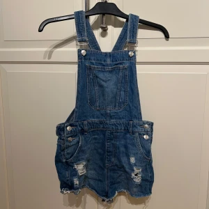  hängselshorts från H&M Divided - Säljer ett par blå jeansshorts med hängslen från H&M Divided. Shortsen har slitna detaljer, fransig kant och klassiska fickor fram och bak. Perfekta för sommaren. 