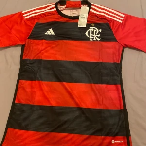 Brasilianska Flamengo tröja  - Säljer en officiell Flamengo fotbollströja från Adidas med klassiska röda och svarta breda ränder. HELT NYA