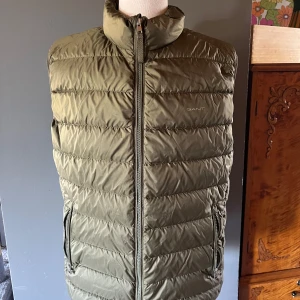 Olivgrön dunväst från Gant - Snygg olivgrön dunväst från Gant med hög krage och dragkedja framtill. Västen har två fickor med dragkedja och quiltad design som ger en sportig look. Perfekt för lager-på-lager under kyliga dagar.