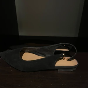 Svarta ballerinaskor med spetsig tå - Säljer ett par svarta ballerinaskor med öppen häl och spetsig tå. Skorna har en tunn rem med spänne runt hälen och är tillverkade i mocka. Perfekta för dig som gillar stilrena och eleganta skor. Har tyvärr ett par likadana 
