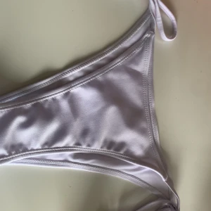 Vit bikiniunderdel med knyt i sidorna - Säljer en stilren vit bikiniunderdel med knyt i sidorna. Klassisk trekantsform och slät yta i syntetmaterial som ger en mjuk och bekväm känsla. Perfekt för sommardagar på stranden eller vid poolen.