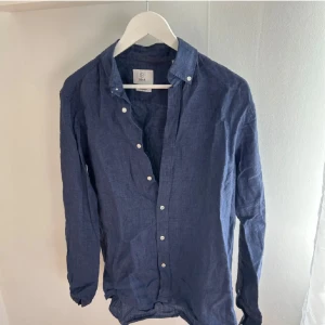 Blå linneskjorta från Bläck - Säljer en stilren blå skjorta i linne från Bläck. Skjortan har lång ärm, klassisk krage och vita knappar framtill. Perfekt för en avslappnad men ändå uppklädd look. Storlek S (37/38).