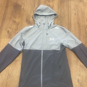 Grå vindjacka med huva från Under Armour - Snygg tvåfärgad grå vindjacka från Under Armour med huva och dragkedja framtill. Jackan har lång ärm, två sidofickor med dragkedja och en sportig look. Perfekt för utomhusaktiviteter när det blåser.