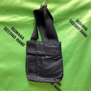 Calvin Klein crossbody väska - Svart crossbody väska från Calvin Klein med bred justerbar axelrem och CK logga längs framsidan. Väskan är tillverkad i syntetmaterial och har en dragkedja upptill.  Mått: Bredd 21 cm, höjd 24 cm   /  Spara skärmtid, besök våra butiker i Sthlm, Malmö & Gbg. New arrivals daily + studentrabatt i alla våra butiker! Hitta till din närmsta butik på humansecondhand.se.   HUMANA Second Hand erbjuder 14 dagars retur, även via plick. Returer skickas med PostNord. Vid retur kontakta support@plick.se.
