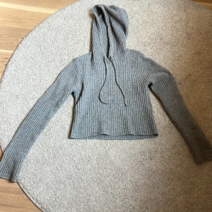 Grå ribbad stickad hoodie - Säljer en grå ribbad stickad hoodie med långa ärmar och huva med dragsko. Tröjan har en mjuk och skön känsla, perfekt för kyligare dagar.
