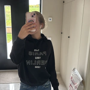 Svart hoodie med tryck - Svart hoodie med vit text på framsidan där det står 'Paris 1985 Berlin 1992'. Tröjan har huva och långa ärmar, samt en klassisk passform. Perfekt för en avslappnad stil. 