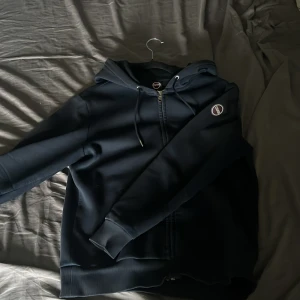 Mörkblå hoodie från Colmar - Säljer en mörkblå hoodie från Colmar med dragkedja och justerbar huva. Tröjan har Colmar-logga på ärmen och ribbade muddar vid ärmslut och nederkant. Perfekt för en avslappnad stil.