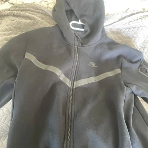 Svart hoodiejacka från Nike - Svart niketeck från JD med dragkedja framtill och reflekterande detaljer över bröstet och på ena ärmen. Jackan har huva och en sportig look, perfekt för träning eller chill. Nike-logga på bröstet.