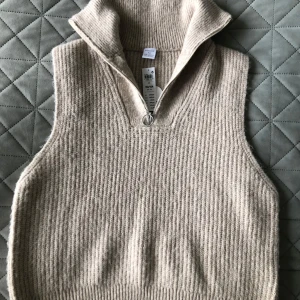 Beige stickad väst/slipover med dragkedja - Beige stickad väst/slipover från Lindex med hög krage och dragkedja framtill. Med ribbad struktur och är ärmlös, perfekt att bära över en skjorta eller tröja för extra värme och stil. Helt ny med lappar kvar på grund av felköp. 
