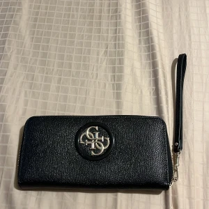 Svart plånbok från Guess - Snygg svart plånbok från Guess med silverfärgad logga framtill och avtagbar handledsrem. Har massa fack och använd cirka 3 gånger bara.