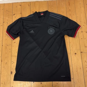 Svart Tyskland fotbollströja från Adidas - Säljer en svart Tyskland fotbollströja från Adidas med diskret klubbmärke och fyra stjärnor på bröstet. Tröjan har korta ärmar med röd och vit detalj vid ärmslut samt klassiska Adidas-ränder på axlarna. Perfekt för fotbollsfans!