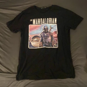 Svart The Mandalorian t-shirt från Star Wars - Säljer en svart t-shirt med tryck från The Mandalorian och Baby Yoda på bröstet. T-shirten har rund hals och korta ärmar. Perfekt för dig som älskar Star Wars och vill ha en snygg och bekväm t-shirt med coolt motiv.