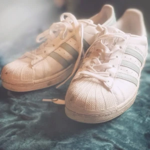 Vita Adidas Superstar sneakers med ljusblå ränder - Säljer ett par klassiska Adidas Superstar sneakers i vitt skinn med ljusblå ränder på sidorna i stl. 38.Skorna har snörning och den ikoniska gummitån framtill. Perfekta för dig som gillar en tidlös och sportig stil.
