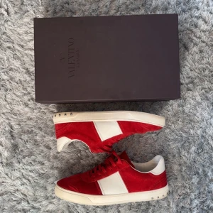 Valentino Garavani röda sneakers med vita detaljer - Säljer ett par röda Valentino Garavani sneakers i mocka med vita skinnpaneler och vita sulor. Skorna har snörning och små silverfärgade nitar vid hälen. Kommer med originalkartong och dustbags. Saknas 1 nit.