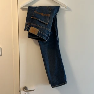 Blå jeans från Nudie Jeans - Säljer ett par klassiska blå jeans från Nudie Jeans med raka ben och snygga kontrastsömmar. Byxorna har fem fickor och en patch med logga bak. Dom är som helt nya och modellen är Lean Dean.Perfekta för dig som gillar stilrena jeans med bra passform. Går att diskutera pris