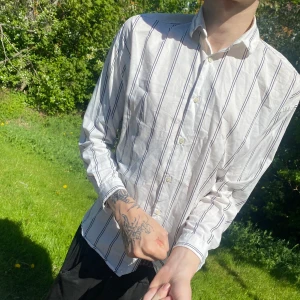 Vit randig skjorta från Zara Man - Säljer en vit skjorta från Zara Man med svarta tunna ränder och relaxed fit. Skjortan har klassisk krage, knäppning framtill och långa ärmar. Passar bra till speciella måltider.