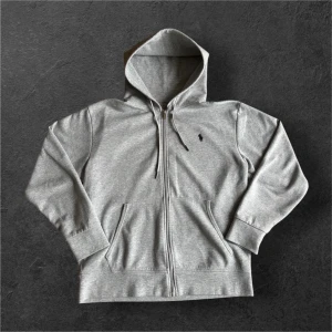 Grå Ralph Lauren hoodie - Klassisk grå hoodie från Polo Ralph Lauren med dragkedja och broderad logga på bröstet. Tröjan har långa ärmar, huva med snörning och två fickor framtill. Perfekt för en avslappnad stil.