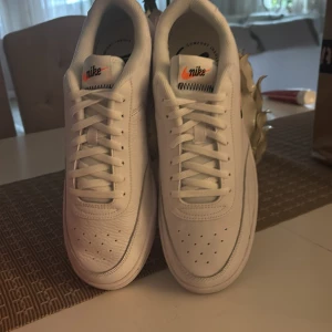 Vita Nike sneakers  - Säljer ett par klassiska vita Nike sneakers med svart och orange logga på tungan. Skorna har snörning, rund tå och är tillverkade i skinn med en platt sula. Perfekta för dig som gillar stilrena och tidlösa sneakers.
