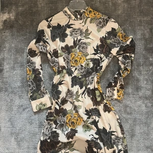 Blommig långklänning från Zara - Långklänning med hög hals från Zara i ett blommigt mönster med gula, grå och svarta toner på en beige botten. Klänningen har långa ärmar och markerad midja för en snygg siluett. Perfekt för dig som gillar statement-plagg med mycket färg och mönster.