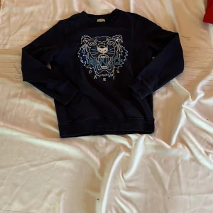 Mörkblå sweatshirt med tiger från Kenzo - Säljer en mörkblå sweatshirt från Kenzo med det ikoniska tigerhuvudet broderat framtill i blå och vita nyanser. Tröjan har rund halsringning och långa ärmar. Perfekt för dig som vill sticka ut med en snygg streetstyle-look. Och såklart äkta