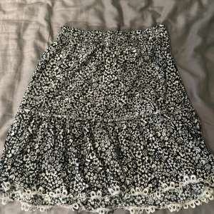 Svartvit blommig kjol från Trendyol - Säljer en svartvit blommig kjol från Trendyol med volang och spetsdetaljer längst ner. Kjolen har elastisk midja och är i kort modell, perfekt för en somrig look.