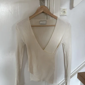 Vit omlottopp från Neo Noir - Säljer en vit omlottopp från Neo Noir i ribbat material. Toppen har lång ärm och djup v-ringning, perfekt för en stilren look. Tillverkad i en mjuk viskos- och nylonblandning. Snygg och enkel att matcha med olika outfits.