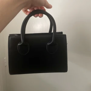 Svart mini handväska - Säljer en stilren svart mini handväska med två handtag. Väskan har en enkel och klassisk design som passar perfekt till att bära det viktigaste. Perfekt accessoar för att lyfta din outfit. Ej använd!! Priset kan diskuteras vid snabb affär 🤗 