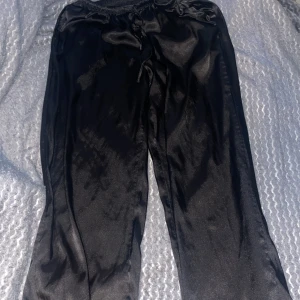 Svarta satinbyxor från hunke möller - Säljer ett par svarta, glansiga satinpyjamasbyxor från hunkemöller  med resår i midjan och dragsko. Byxorna har en loose passform och är perfekta för en avslappnad men ändå stilren look. Endast använda en gång.