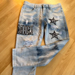 Ljusblå jeans med stjärnor och broderad text från Amiri - Unika ljusblå jeans med slitningar, broderade svarta stjärnor och stor svartvit Amiri-text på ena benet. Jeansen har en tvättad look och raka ben. Perfekta för dig som vill sticka ut med detaljer och cool design.