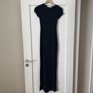Svart långklänning från Bershka - En stilren svart långklänning från Bershka med rund halsringning och korta ärmar. Klänningen har en tight passform och är perfekt för dig som gillar minimalistisk stil. Enkel att styla med sneakers eller klackar.