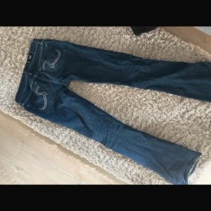 Blå bootcut jeans från Replay - Säljer ett par klassiska blå bootcut jeans från rock and republic med snygga broderade bakfickor i lila och vitt. Jeansen har normal passform och är tillverkade i jeansmaterial. Perfekta för dig som gillar en tidlös stil.