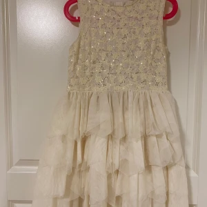 Vit ärmlös klänning med volanger och paljetter - Superfin vit klänning med ärmlös design, glittrig överdel med paljetter och spets, samt flera lager volanger i kjolen. Perfekt för speciella tillfällen när du vill sticka ut lite extra.