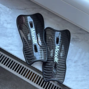 Monolit benskydd i kolfiber - Säljer ett par svarta benskydd från Monolit i kolfiber med vit logga. De är lätta och har en snygg glansig finish med kolfibermönster. Insidan är vadderad för extra komfort och skydd vid spel.