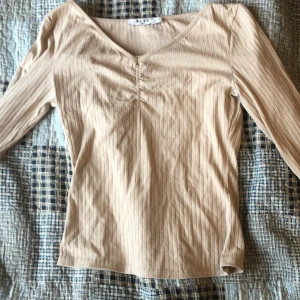 Beige ribbad topp från NA-KD - Säljer en beige långärmad topp från NA-KD med v-ringning och ribbad struktur. Toppen har en lätt rynkning vid bysten och är figurnära i modellen. Perfekt för lager-på-lager eller som den är.