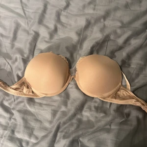 Beige bh från H&M - Säljer en beige bh från H&M med släta kupor och lätt vaddering. Bh:n har en enkel och stilren design med tunna axelband och diskreta spetsdetaljer vid kuporna. Perfekt under ljusa toppar.