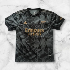 Arsenal svart bortatröja 2022/23 - Snygg svart Arsenal bortatröja från Adidas med guldiga detaljer och loggor. Tröjan har geometriskt mönster och korta ärmar. Perfekt för fotbollsfans som vill sticka ut på läktaren eller planen.