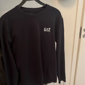 Svart långärmad tröja från EA7 Emporio Armani - Snygg svart långärmad tröja från EA7 Emporio Armani med vit logga på bröstet och diskret detalj på ryggen. Perfekt för en stilren och sportig look. Rund halsringning och mjukt material.