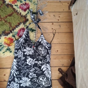 Svartvit blommig halterneckklänning - Snygg svart klänning med vita blommor och blad, halterneck med knyt i nacken och smala axelband. Perfekt för varma dagar och somriga tillfällen.