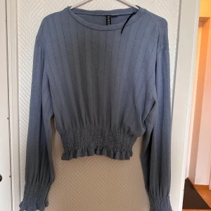 Ljusblå ribbad tröja från Divided - Säljer en ljusblå ribbad tröja från Divided med smockad midja och ärmslut. Tröjan har långa ärmar och en rund halsringning. Perfekt för dig som gillar en feminin och trendig stil.