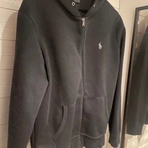Svart hoodie från Ralph Lauren - Säljer en stilren svart hoodie från Ralph Lauren med dragkedja och det klassiska logotypbroderiet på bröstet. Tröjan saknar snören på luvvan 