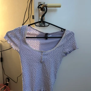 Ljuslila topp med småblommigt mönster - Söt ljuslila topp med små vita blommor och rundad urringning. Toppen har korta ärmar och tre dekorativa knappar framtill. Perfekt för en avslappnad och trendig look.