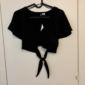 Svart croppad knytblus från H&M Divided - Säljer en svart croppad blus från H&M Divided med korta ärmar och knytning baktill. 