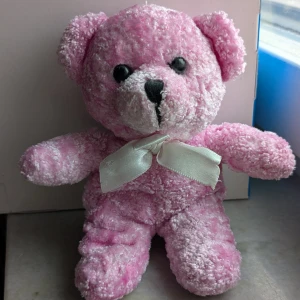Rosa nallebjörn med vit rosett - Söt och fluffig rosa nallebjörn med en vit rosett runt halsen. Perfekt som present eller dekoration. Mjuk och kramvänlig!