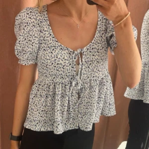 Blommig blus med knytning - Charmig blommig blus i vitt och blått med korta puffärmar och knytning framtill. Perfekt för en somrig look.